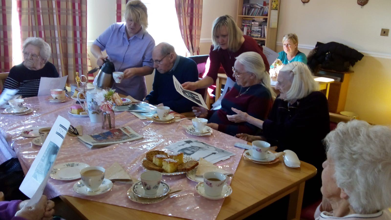 A picture of several older white people sitting around a table with tea and cakes