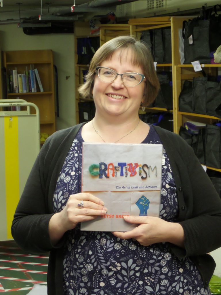 Picture of a smiling white woman with glasses, looking at the camera and holding a book with the word 'Craftivism'